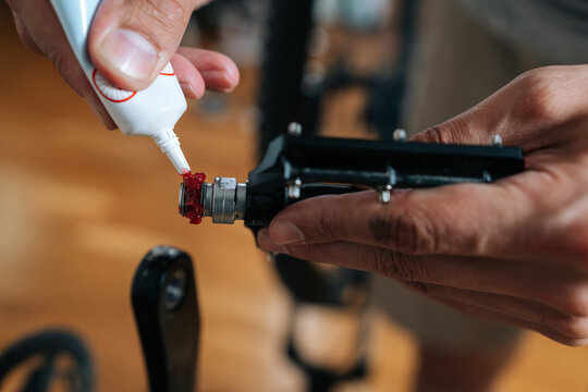 Close-up hand of unrecognizable person applying red grease from tube onto bicycle pedal bearing, performing essential maintenance for smooth cycling performance and component longevity.