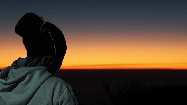 Silhouetted figure watches sunrise from hilltop. Orange glow meets dark sky in tranquil moment. Cool tones contrast with warm horizon light. Quiet observer enjoys peaceful dawn arrival