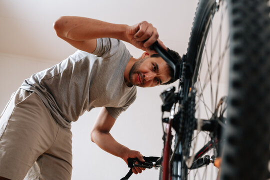 Low-angle view of man with concentrated expression works on bicycle indoors. DIY bike maintenance and repair concept. Casual lifestyle scene of cyclist fixing own equipment in house, apartment.