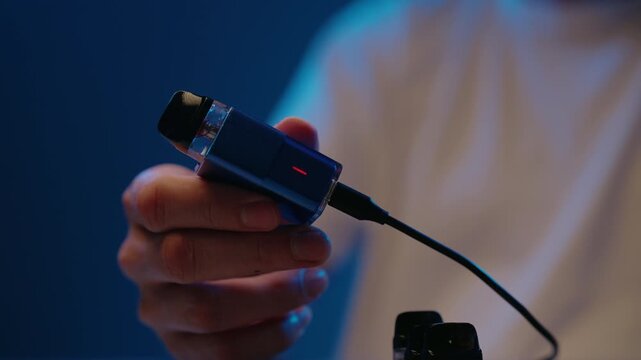 Person plugging usb cable into blue vape pen for charging, connecting to mobile phone with reverse charge capability for power, with green light indicating charging, close-up, slow motion.