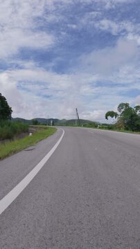 Scenic mountain road curving through lush green countryside, with distant hills, utility poles and a blue sky filled with soft clouds.