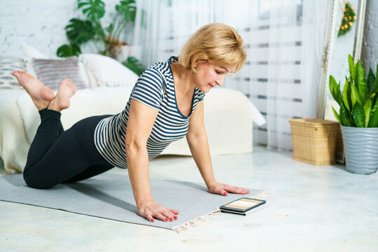 Mature female in striped shirt holding kneeling plank on mat in bedroom, watching online video guide on smartphone. Core training and active aging at home.