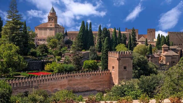 Breathtaking view of Gardens and Architecture of the Alhambra Palace in Granada, Spain. The historic Moorish red-stone fortifications, towers, and bell tower rise above surrounding green trees and gar