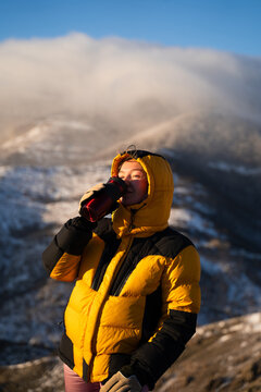 Woman enjoying hot drink from thermos cup in winter mountains at sunrise, wearing warm down jacket, outdoor lifestyle, adventure and travel concept.