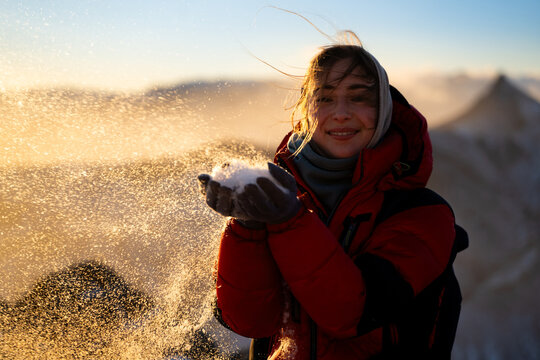 Smiling candid woman playing with snow in golden sunrise light on mountain summit in winter season, feeling happiness, freedom and enjoying outdoor lifestyle concept.