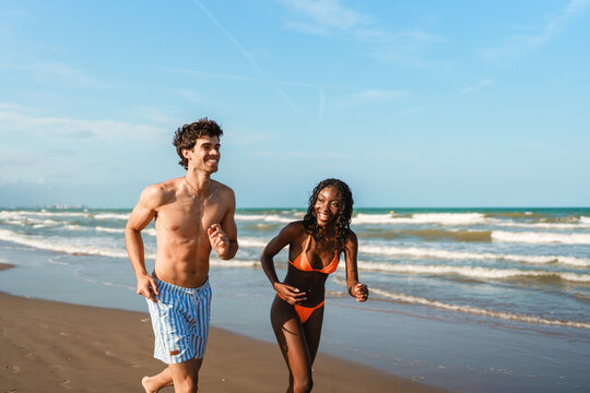 Happy young couple jogging on sandy beach beside ocean waves under blue sky
