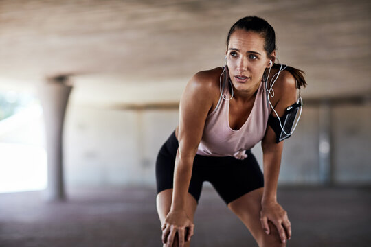 Earphones, woman and tired in city for fitness, catch breath and exercise break with streaming music. Parking lot, runner or reflection with tech for wellness, workout fatigue or cardio tips in audio