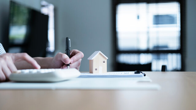 Home buyer signing documents and calculating housing costs with a wooden house model, concept of mortgage, real estate purchase, financial planning and property investment.