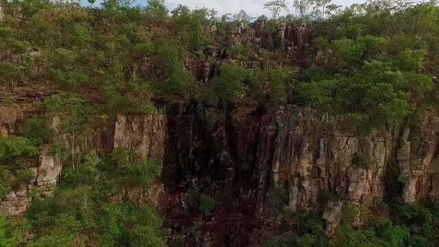 Quartzite cliff rises above dense Cerrado shrubland at Almecegas, with weathered rock faces, native vegetation and rolling uplands near Alto Paraiso de Goias, drone ascent