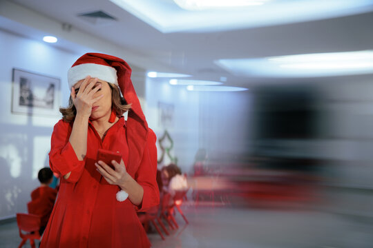 Frustrated Woman In Santa Hat Looking At Smartphone Screen Display. Disappointed lady holding red mobile phone makes facepalm gesture during winter holiday celebration