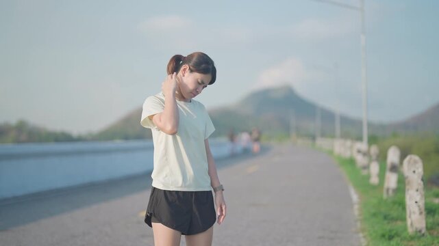 Young Asian fitness woman holding her injured shoulder and neck due to muscle pain during training. Runner female having body problem after outdoor exercise in summer. Sports and medical concept