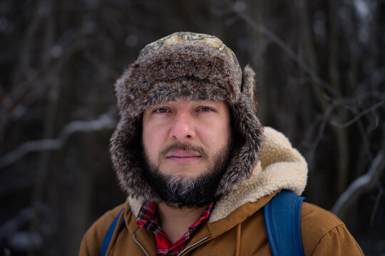 Serious bearded man in fur trapper hat in winter forest