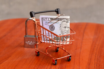Miniature shopping cart filled with US dollar banknotes next to a steel padlock on a wooden table, secure online shopping and financial data protection concept. © Isman