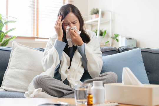 Sick Asian woman blowing her nose with tissue on sofa, wrapped in blanket, suffering from high fever, seasonal flu, influenza virus infection, and cold symptoms at home