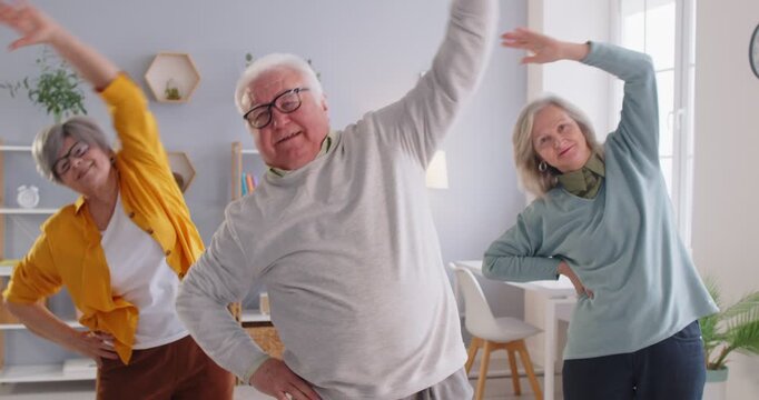 Group of happy senior retired people doing exercises in retirement home, leaning to sides during morning gymnastics. Smiling pensioners stretching and staying active to maintain health and mobility.