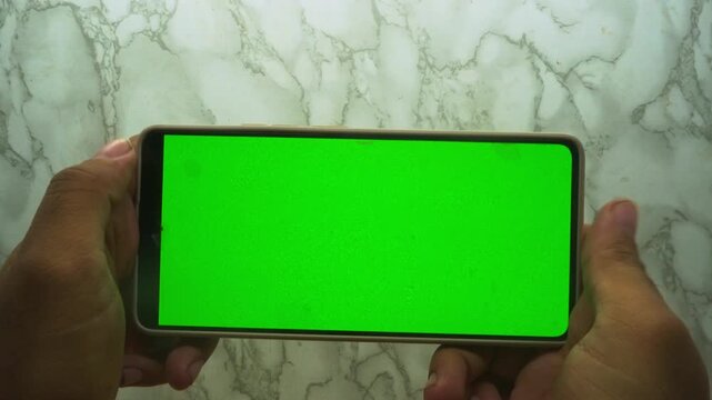 Hands holding a smartphone with a blank green screen display against a textured white marble surface background for mobile content apps mockup presentation and digital media marketing design