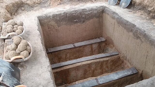 A prepared Muslim burial grave designed with three separate sections or compartments for the burial of three deceased persons, with wooden supports, soil-filled baskets, and shovels surrounding the gr