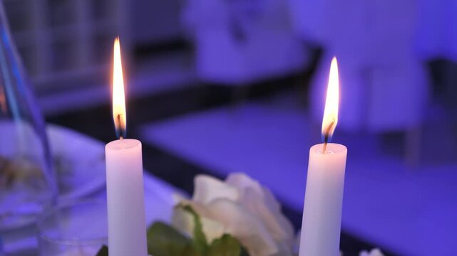 Close-up of two white wax candles burning with steady flames on a festive table with blue atmospheric lighting and blurred background.