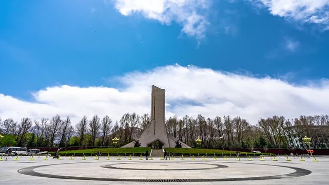 Tibetan Monument in Lhasa Square under blue sky with scattered clouds, wide-angle static shot showing paved plaza, circular pattern, bare trees, and distant mountains
