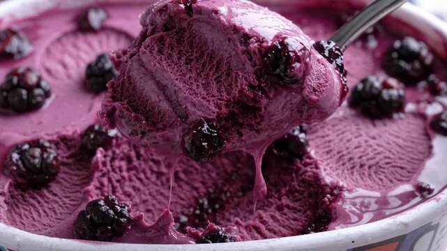Close view of metallic spoon lifting generous scoop of purple blackberry frozen dessert from container, revealing creamy texture, icy granules, glossy berry glaze, and slow dripping sweetness.