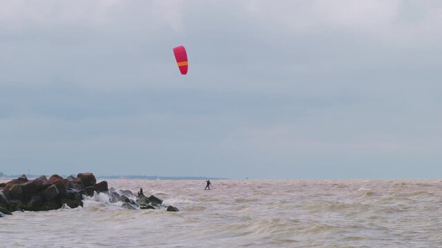 Action wide shot of a lone kitesurfer with a red kite gliding across rough ocean waters on an overcast day. Extreme water sports and active lifestyle concept. Flying over water in slow motion..