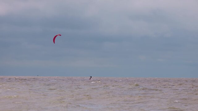Action wide shot of a lone kitesurfer with a red kite gliding across rough ocean waters on an overcast day. Extreme water sports and active lifestyle concept. Flying over water in slow motion..