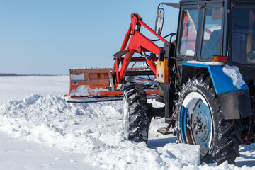 Tractor with snow plow stands in snowy field. Bright sun shines overhead on clear day.