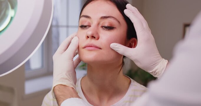 Doctor cosmetologist or beautician examining skin condition for female client with special lamp in beauty medical clinic. Cosmetology, facial treatment and beauty procedure concept. 4k video.