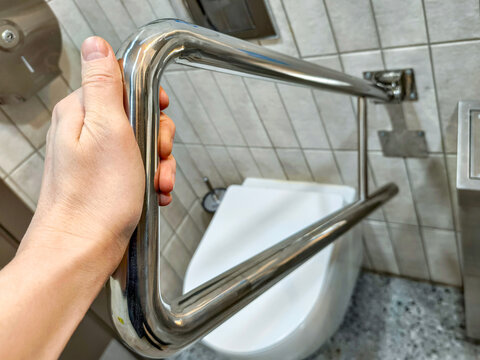Hand of man holding stainless steel grab bar in accessible restroom. Support rail for disabled person in public toilet
