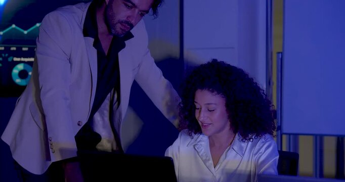 Adult female professional reviews laptop data while male manager leans in, points toward screen, and exchanges ideas about performance dashboard metrics during collaborative business analysis.