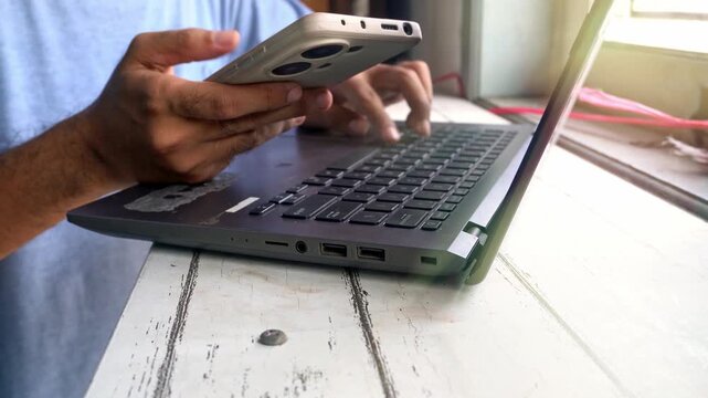 A close up view of a busy person multitasking by using a modern smartphone while working on a laptop computer in a well lit office environment during a normal productive workday at the desk