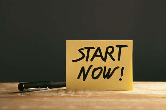 Start now message on sticky note with pen on wooden table and black background