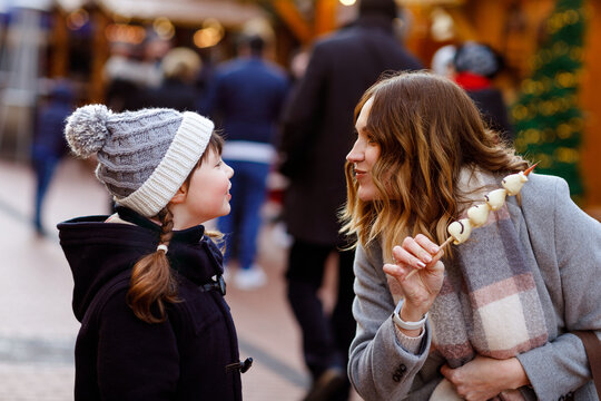 Young mother and daughter eating white chocolate-covered fruits and strawberries on skewers at German Christmas market. Snowy festive fun. Real life, real people, family moment