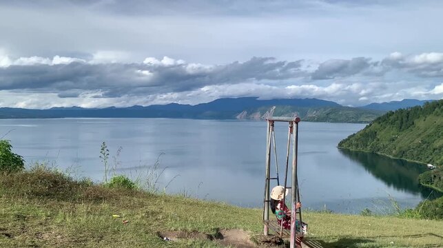 Stock footage of a young woman sitting on a metal swing with a wide scenic backdrop of a calm lake and mountains at Tara Bunga Hill, North Sumatera, Indonesia under cloudy blue sky. Travel theme.