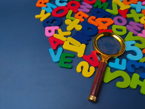 Gold brass magnifying glass placed over scattered colorful wooden letters and numbers against a dark blue background