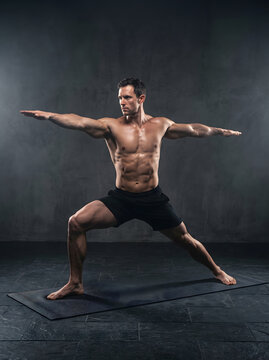 A powerful shot of a muscular man performing the Warrior II yoga pose on a mat in a dimly lit studio. This image captures dedication, balance, and the pursuit of a healthy, active lifestyle through...