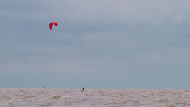 Action wide shot of a lone kitesurfer with a red kite gliding across rough ocean waters on an overcast day. Extreme water sports and active lifestyle concept. Flying over water in slow motion..