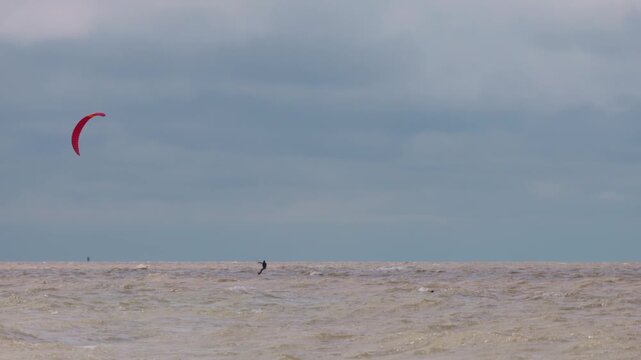 Action wide shot of a lone kitesurfer with a red kite gliding across rough ocean waters on an overcast day. Extreme water sports and active lifestyle concept. Flying over water in slow motion..