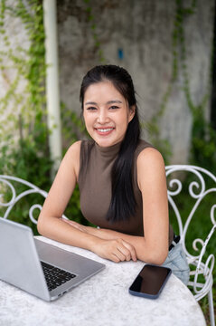 Portrait Of Smiling Asian Woman Sitting Beside Laptop Outdoors