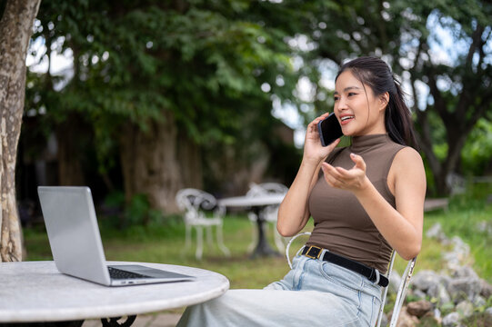 Asian Woman Making Phone Call Beside Laptop In Outdoor Garden