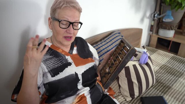 Counting on a wooden abacus. Senior woman talking and writing in notebook while sitting on bed.