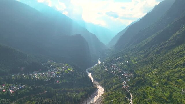 Aerial establishing shot gliding over Barshaini village in the deep Parvati Valley, revealing dramatic sunbeams piercing monsoon clouds in Kullu District, India.