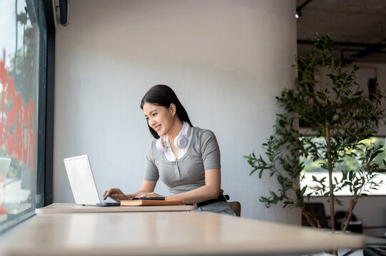 Young Asian woman with headphone around neck using laptop while studying at table in co working cafe