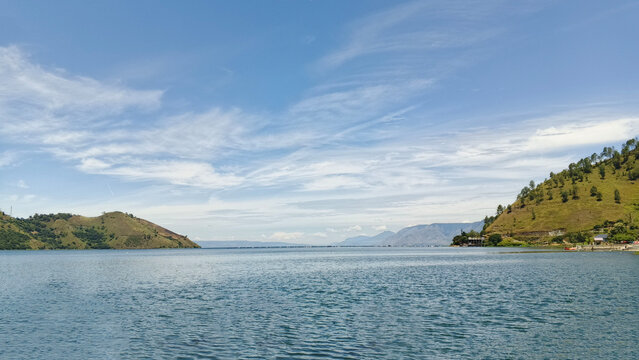 view of Lake Toba with calm blue waters, surrounding green hills, and a clear blue sky with light clouds.