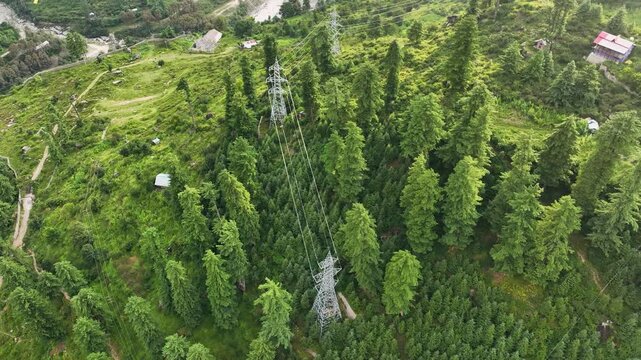 High angle drone tracking shot along high-voltage transmission towers and power lines cutting across lush pine forests in Parvati Valley, Himachal Pradesh.