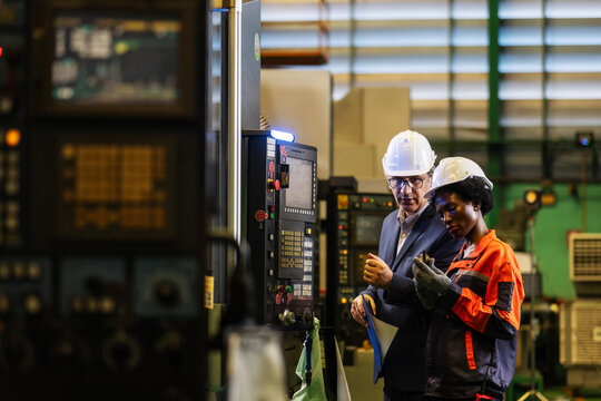 Male and female professionals discuss CNC machine operation beside a control panel in a factory. Wearing safety helmets, they participate in practical workplace training