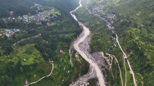 Aerial establishing shot gliding over Barshaini village in the deep Parvati Valley, revealing dramatic sunbeams piercing monsoon clouds in Kullu District, India.