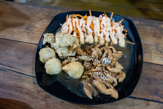 Close up shot of sliced chicken teriyaki topped with sesame seeds, fresh carrot salad with mayonnaise, and deep fried side dishes served on a dark plate over a rustic wooden table.