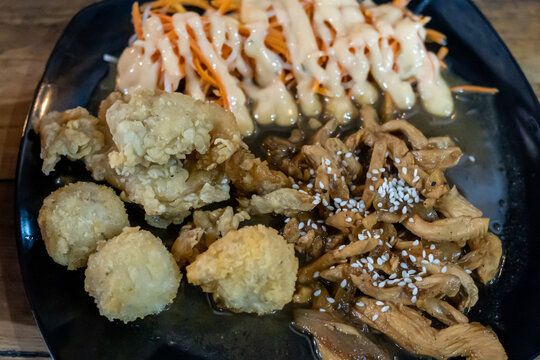Extreme close up shot of chicken teriyaki topped with sesame seeds, creamy shredded carrot salad with mayonnaise, and deep fried snacks served on a dark plate.