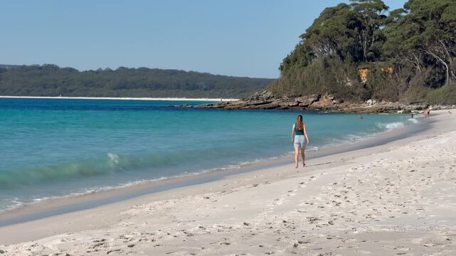 A relaxed girl walks along the white sand of Chinamans Beach, with turquoise water, gentle waves and surrounding bush, near Wollongong, Jervis Bay, New South Wales, Australia.
